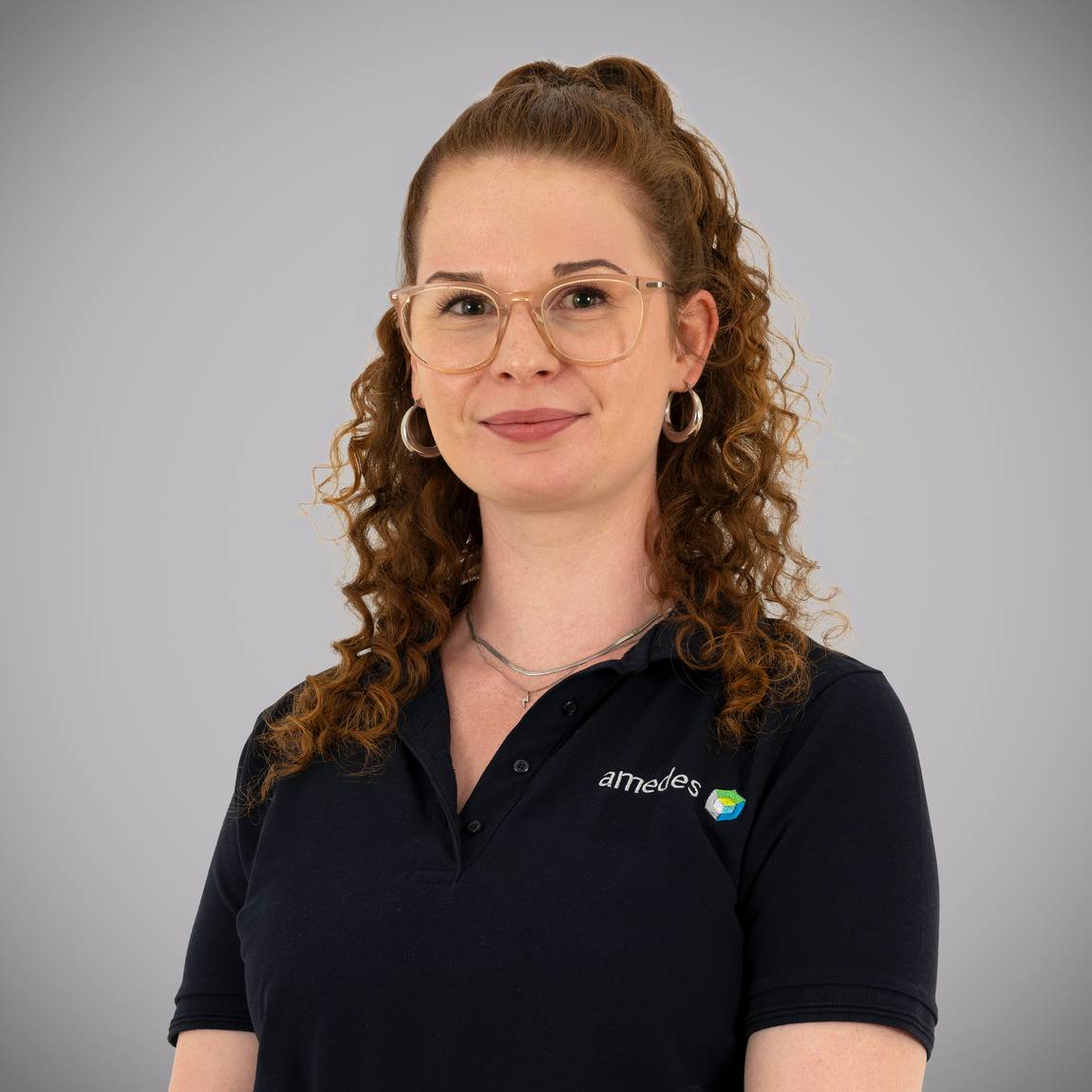 Eine Frau mit lockigem kastanienbraunem Haar, die eine Brille und Ohrringe trägt, lächelt leicht. Sie trägt ein marineblaues Poloshirt mit dem aufgestickten amedes-Logo. Der Hintergrund ist einfarbig grau.
