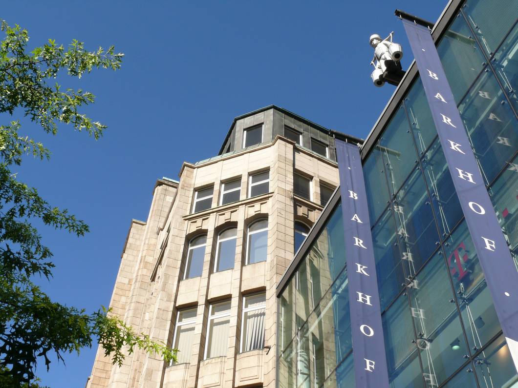 Ein modernes Gebäude aus Glas und Stein mit blauen BARKHOF-Bannern und einer weißen Statue einer sitzenden Figur, die hoch an der Ecke angebracht ist, vor einem klaren blauen Himmel mit einem Baumzweig auf der linken Seite.