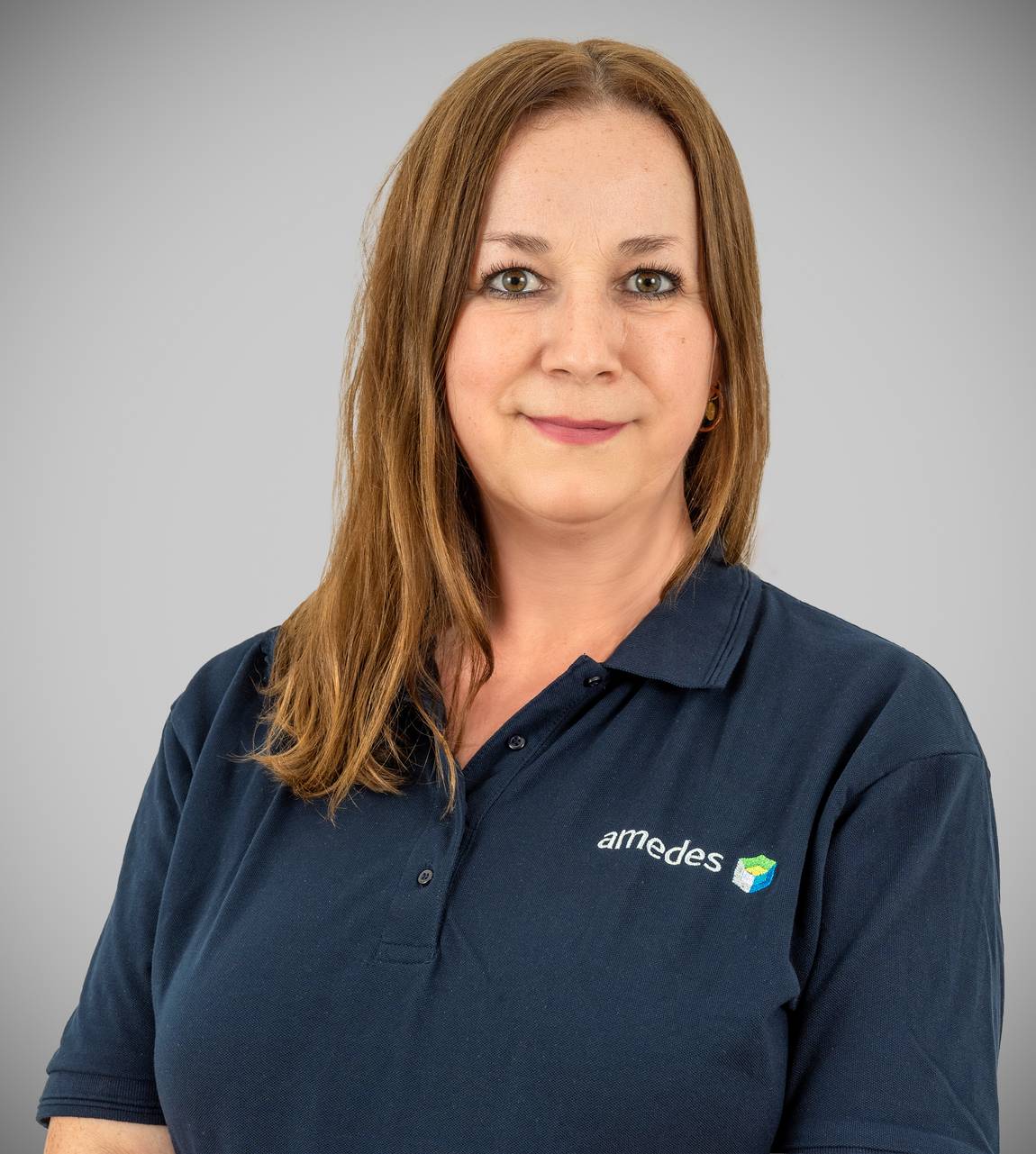 Eine Frau mit glattem, hellbraunem Haar, die ein marineblaues Poloshirt mit einem amedes-Logo trägt, steht vor einem schlichten grauen Hintergrund und lächelt sanft in die Kamera.