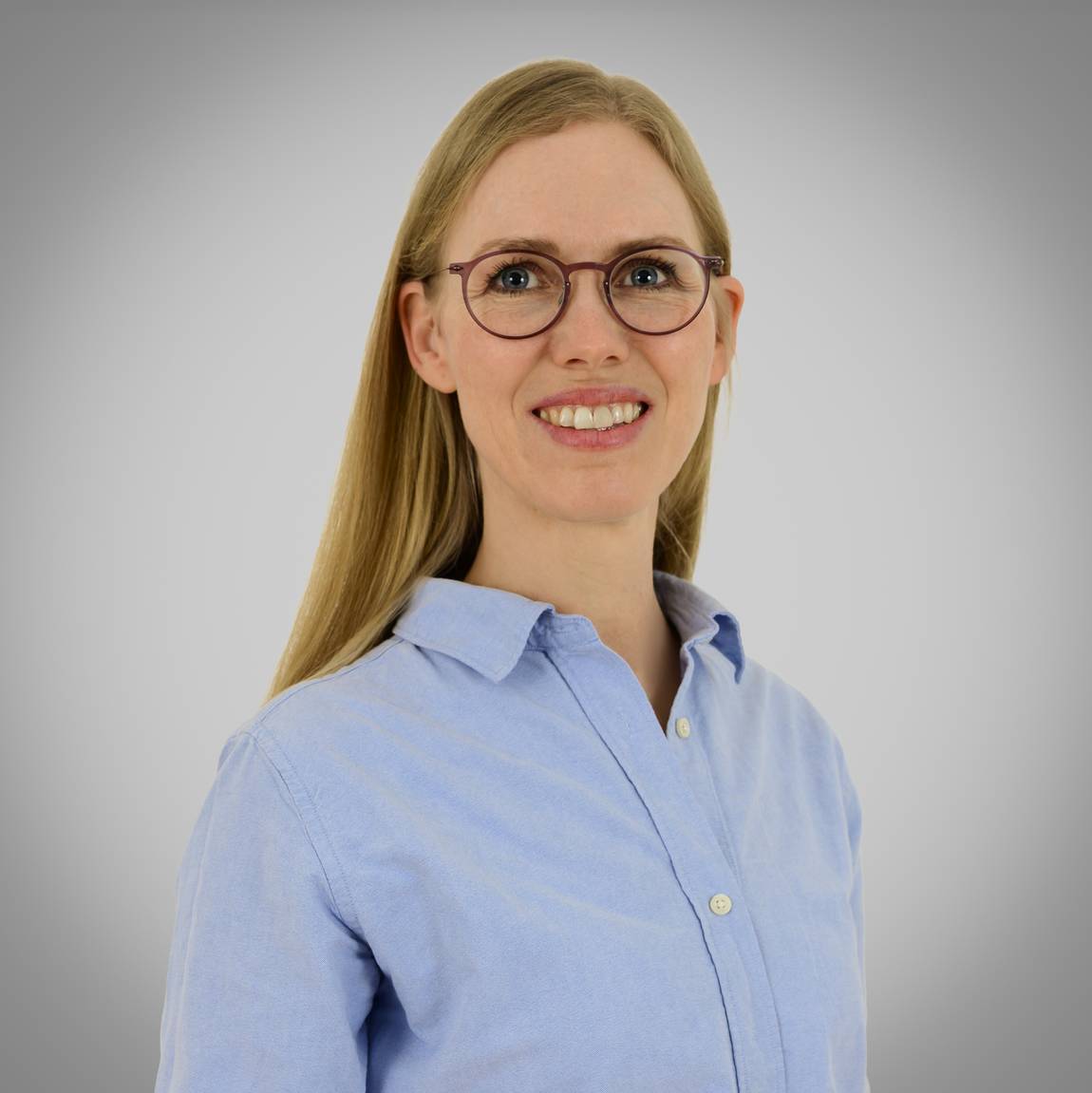 Eine Frau mit langen blonden Haaren, die eine Brille und ein hellblaues Button-up-Hemd trägt, steht lächelnd vor einem schlichten grauen Hintergrund.