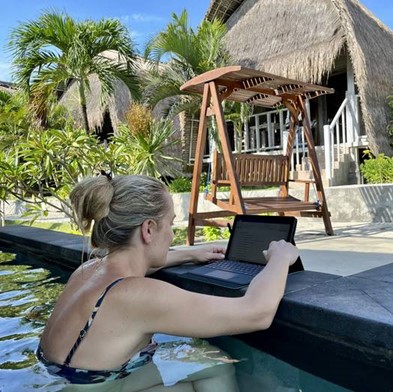 Eine Frau in einem Pool arbeitet an einem Laptop, der auf dem Beckenrand steht. Im Hintergrund sind tropische Pflanzen, Palmen, eine strohgedeckte Cabana und eine Holzschaukel unter blauem Himmel zu sehen.